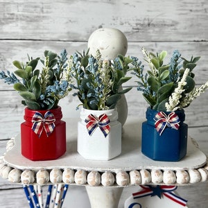 Pode incluir: Um suporte branco de v&aacute;rios n&iacute;veis com decora&ccedil;&otilde;es vermelhas, brancas e azuis. O n&iacute;vel superior tem tr&ecirc;s pequenos frascos com vegeta&ccedil;&atilde;o e la&ccedil;os vermelhos, brancos e azuis. O n&iacute;vel intermedi&aacute;rio tem um frasco vermelho com canudos. O n&iacute;vel inferior tem uma placa que diz "my home sweet home" e um buqu&ecirc; de rosas vermelhas com vegeta&ccedil;&atilde;o.
