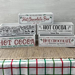 May include: Four white wooden boxes stacked on top of each other. Each box has a different design with the words "Hot Cocoa" or "Hot Chocolate" in red and green lettering. The top box says "The Pollack Family Hot Chocolate Bar Warm Up Here Open All Winter est 2012". The second box says "Warm Up Here Hot Cocoa Served Here Est 1850". The third box says "Hot Cocoa Marshmallows Candy Canes Cookies Served Here". The bottom box says "Old Fashioned Hot Chocolate Warm Up Here Since 1854".