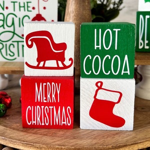 Peut inclure: Un plateau en bois à étages avec un bord perlé, décoré de blocs en bois sur le thème de Noël. Les blocs présentent des phrases comme "Croire en la magie", "Chocolat chaud", "Joyeux Noël" et "Croire". Il y a aussi des blocs avec des images d'un traîneau, d'une chaussette et d'une branche de houx.