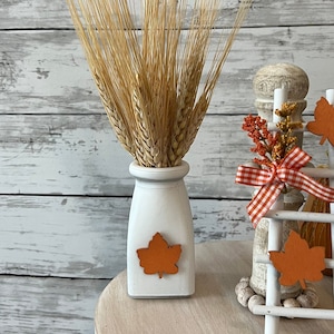 May include: A white ceramic vase with a fall leaf design holds a bunch of dried wheat stalks. A white wooden ladder with fall leaf accents sits on a white table with a wooden top.
