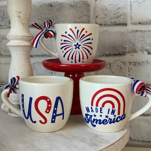 May include: Three white ceramic mugs with red, white, and blue patriotic designs. The mugs are on a white tiered cake stand. The mugs feature the words "USA", "Made in America", and a firework design.