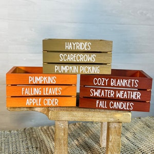 May include: Three wooden crates stacked on a small wooden stool. The top crate is brown and says "HAYRIDES, SCARECROWS, PUMPKIN PICKING". The middle crate is orange and says "PUMPKINS, FALLING LEAVES, APPLE CIDER". The bottom crate is red and says "COZY BLANKETS, SWEATER WEATHER, FALL CANDLES".