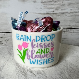 May include: White ceramic mug with a pink, purple, and green floral design and the text "Rain Drop Kisses and Flower Wishes". The mug is filled with colorful chocolate candies.