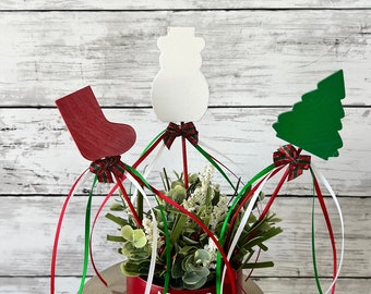 May include: A red pot with the words "Merry Christmas" on it, decorated with a snowman, a stocking, and a Christmas tree. The pot is surrounded by green, red, and white ribbon.