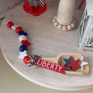 May include: A wooden scoop with the word "LIBERTY" painted on it. The scoop is filled with red, white, and blue stars. The scoop is sitting on a white tiered tray with a red, white, and blue beaded garland.