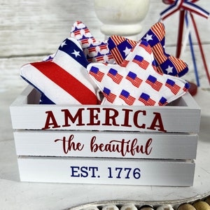 May include: A white wooden crate with the words "AMERICA the beautiful EST. 1776" printed on the front. The crate is filled with red, white, and blue fabric stars.