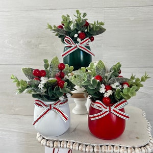 May include: Three small potted Christmas arrangements with red berries, green leaves, and white faux snow. The pots are painted green, white, and red and have a red and white ribbon tied around them. The pots are sitting on a white tiered stand with a sign that says "hello christmas" and another sign that says "santa mistletoe hot cocoa snow".