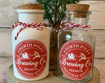 Könnte beinhalten: Zwei weiße Glasbehälter mit Korkdeckeln und roter und weißer Schnur, die um die Hälse gebunden sind. Die Behälter sind mit einem roten Kreis mit weißem Text verziert, der "North Pole Brewing Co. Est. 1824 Premium Christmas Spirits" lautet.