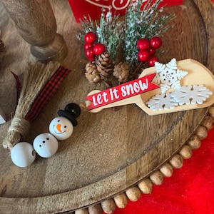 May include: A wooden tiered tray with a red sign that says "It's the Time of the Year" and a wooden scoop with the words "Let it Snow" and white snowflakes. There are three white snowman beads and a brown and red tassel on the tray.