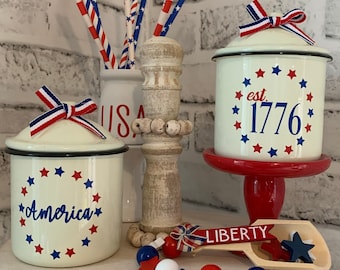 May include: A tiered white cake stand with red, white, and blue decorations. The top tier has a white ceramic jar with a blue and red star design and the text "America". The middle tier has a white ceramic jar with a blue and red star design and the text "est. 1776". The bottom tier has a wooden scoop with the text "LIBERTY" and a string of red, white, and blue beads.
