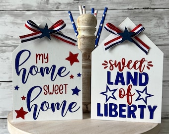 May include: Two white wooden signs with red, white, and blue stars and ribbon accents. The signs read "My Home Sweet Home" and "Sweet Land of Liberty".