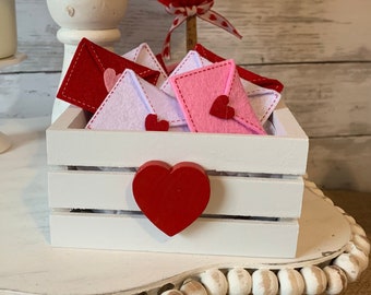May include: White tiered cake stand with a white wooden crate filled with pink and white felt envelopes. The crate has a red felt heart on the front. A red felt heart is on a stick in the background.