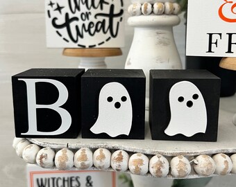 May include: Black and white wooden blocks spelling "BOO" with a ghost on each of the "O" blocks. The blocks are on a white tiered stand with white beads around the edges. There are other Halloween decorations on the stand, including a sign that says "trick or treat" and a sign that says "Happy Halloween".