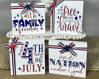 May include: Four wooden blocks with red, white, and blue patriotic decorations and text. The blocks read "faith family freedom", "home of the free because of the brave", "happy 4th of july", and "one nation under god".