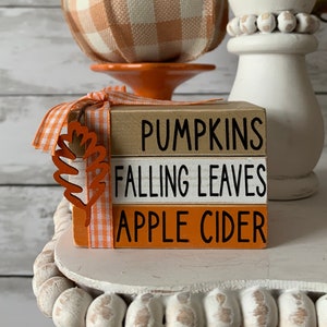 May include: A set of three wooden blocks stacked on top of each other. The blocks are painted in orange, white, and brown. The top block reads "Pumpkins", the middle block reads "Falling Leaves", and the bottom block reads "Apple Cider". The blocks are tied together with a white and orange checkered ribbon and a small orange leaf.