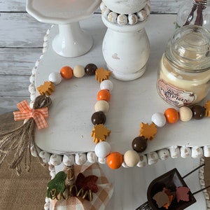 Pode incluir: Um suporte branco de vários níveis decorado com itens temáticos de outono. O nível superior apresenta uma placa que diz "Farm Fresh Pumpkins - Picked Daily". O nível intermediário apresenta um tampão de garrafa de cerâmica branca, uma vela com um rótulo "White Pumpkin Vanilla" e uma guirlanda de contas laranja, marrom e branca com detalhes de folhas de madeira. O nível inferior apresenta uma pequena abóbora xadrez e uma carriola em miniatura com folhas.