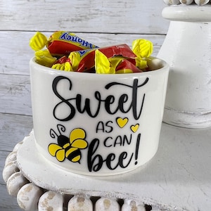 May include: White ceramic jar with a black and yellow bee graphic and the text "Sweet as can bee!"  The jar is filled with yellow and red wrapped candies.