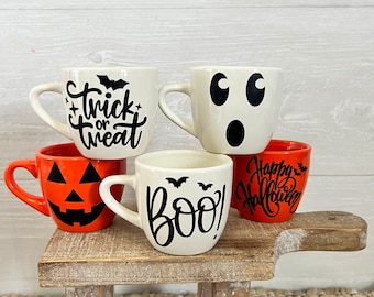 Puede incluir: Conjunto de cuatro tazas de cerámica con temática de Halloween. Dos tazas son blancas con letras y diseños negros, una dice "trick or treat" con un murciélago y la otra dice "boo!" con dos murciélagos. Dos tazas son naranjas con letras y diseños negros, una dice "Happy Halloween" con un murciélago y la otra tiene una cara de calabaza.