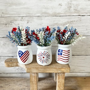 May include: Three white ceramic jars with red, white, and blue patriotic designs. Each jar is filled with a bouquet of blue and white flowers with red accents. The jars are sitting on a small wooden stool.