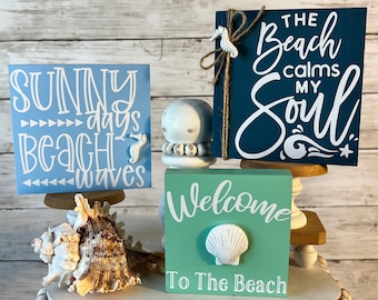 May include: Three wooden signs with beach-themed sayings. The top sign is dark blue with white text that reads "The Beach Calms My Soul". The middle sign is light blue with white text that reads "Sunny Days Beach Waves". The bottom sign is light green with white text that reads "Welcome To The Beach".
