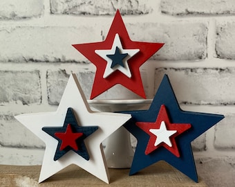 May include: Three wooden star decorations in red, white, and blue, arranged on a small wooden table. Each star has a layered design with smaller stars nested inside. The background features a white brick wall.