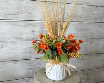 May include: A white ceramic vase with a rustic finish, filled with a floral arrangement of orange flowers and green leaves. The vase is topped with wheat stalks and sits on a weathered wooden stool.