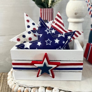 May include: A white wooden crate with red, white, and blue ribbon and a large red, white, and blue star. The crate is filled with blue, white, and red fabric stars.
