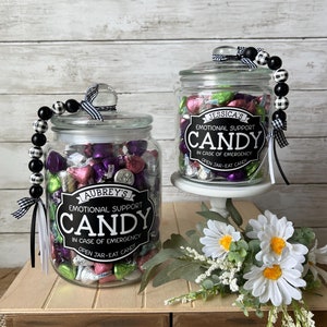 May include: Two glass jars filled with wrapped candies, labeled "Emotional Support Candy." One jar is on a wooden crate, the other on a white pedestal. Each jar has a decorative beaded garland and a black and white ribbon.