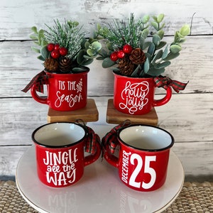 May include: Four red ceramic mugs with black rims and white Christmas-themed text. The mugs are decorated with sprigs of greenery, red berries, and pine cones. Two of the mugs are on a wooden stand, and the other two are on a white cake stand. The text on the mugs reads "Tis the Season", "Holly Jolly", "Jingle All the Way", and "December 25".