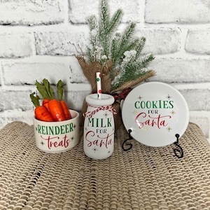 May include: Three ceramic containers with festive Christmas designs. The containers are white with green and red lettering. The first container says "Reindeer Treats", the second says "Milk for Santa", and the third says "Cookies for Santa".