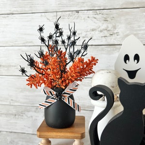 Pode incluir: Uma bandeja de três níveis para Halloween decorada com detalhes pretos e laranja. A bandeja apresenta uma figura de gato preto, uma figura de fantasma, um vaso preto com galhos de aranha laranja e preto, e blocos de madeira com designs temáticos de Halloween.