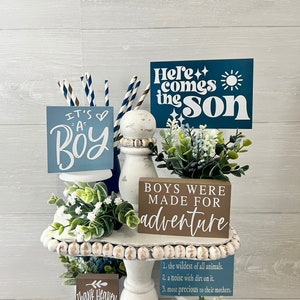 May include: A tiered white stand with three signs. The top sign is blue with white text that reads "Here comes the son". The middle sign is brown with white text that reads "Boys were made for adventure". The bottom sign is brown with white text that reads "Thank Heaven for little boys".