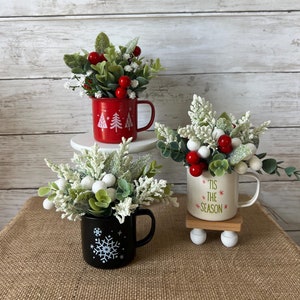 May include: Three Christmas-themed mugs with greenery and red berries. The mugs are decorated with snowflakes, Christmas trees, and the text "Tis the Season".