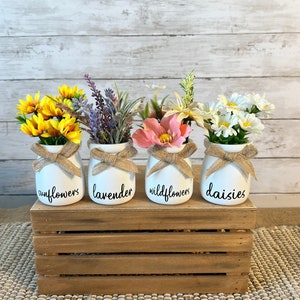 May include: Four white ceramic jars with burlap ribbon and handwritten labels: "sunflowers", "lavender", "wildflowers", and "daisies". Each jar contains a different type of artificial flower arrangement. The jars are displayed on a wooden crate.