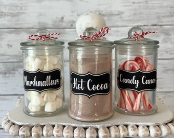 May include: Three glass jars with black chalkboard labels on a white tiered stand. The labels read "Marshmallows", "Hot Cocoa", and "Candy Canes". The jars are filled with white marshmallows, brown cocoa powder, and red and white candy canes.
