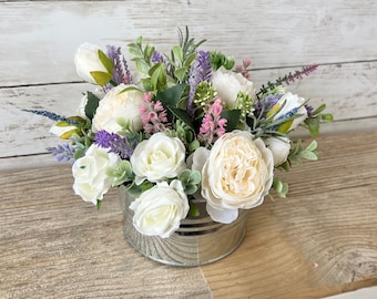 May include: A white and cream floral arrangement in a silver metal container. The arrangement includes white roses, lavender, and greenery.