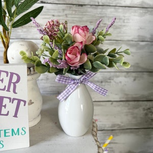 May include: A white ceramic vase with a purple and white checkered ribbon around the neck. The vase is filled with a bouquet of pink roses and purple flowers.