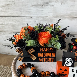 Pode incluir: Uma caixa de madeira preta com uma fita xadrez preta e laranja e o texto "Happy Halloween Witches". A caixa está cheia de um arranjo floral de flores laranja e pretas, folhas verdes e bagas pretas. A caixa está sentada em uma bandeja branca com contas pretas e laranja.