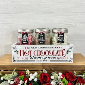 May include: A white wooden box with the text "Old Fashioned Hot Chocolate Warm up here since 1834" on the front. The box contains three glass jars filled with candy canes, hot cocoa mix, and marshmallows.