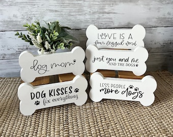 May include: Four white wooden bone-shaped signs with black text. The signs read "dog mom", "DOG KISSES fix everything", "LOVE IS A four legged word", and "just you and me AND THE DOGS" and "LESS PEOPLE more dogs".