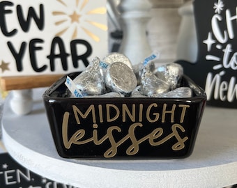 May include: A black ceramic dish with gold lettering that reads "MIDNIGHT KISSES". The dish is filled with silver foil-wrapped chocolate kisses.