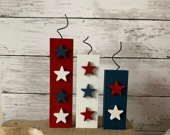 May include: Three wooden blocks painted in red, white, and blue with white and blue stars. The blocks are arranged in a row on a wooden stool. The blocks are decorated with a black wire that resembles a fuse.