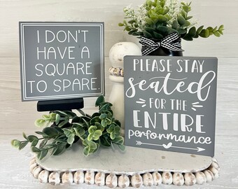 May include: Two gray wooden signs with white text. The first sign says "I don't have a square to spare." The second sign says "Please stay seated - for the entire performance." Both signs are on a white pedestal with a small green plant on each side.