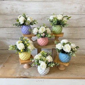 May include: Six small white flower arrangements in pastel colored ceramic pots on a tiered wooden stand. The pots have a geometric pattern.