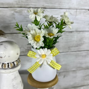 May include: White ceramic egg-shaped vase with a yellow and white gingham ribbon bow and a white daisy on top. The vase is filled with a bouquet of white daisies with yellow centers.