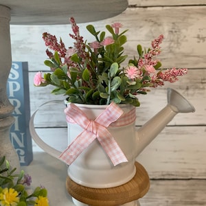 May include: A white watering can filled with pink flowers and greenery, adorned with a pink and white gingham ribbon. The watering can sits on a small wooden base with white feet, part of a tiered display. The word "SPRING" is visible in the background.