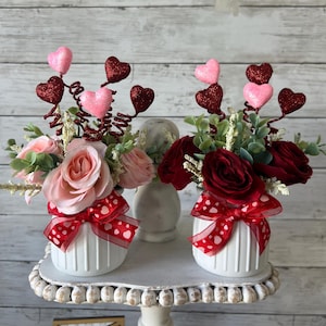 May include: Two white ceramic vases with red and white polka dot ribbon bows. Each vase is filled with artificial flowers, one with pink roses and the other with red roses. Both vases are topped with red and pink glitter hearts on wire stems.