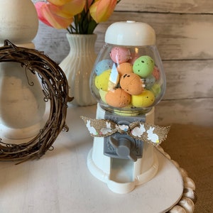 May include: White ceramic Easter egg gumball machine with a clear plastic container filled with pastel colored Easter eggs. The machine has a gray crank and a burlap bow with a white bunny rabbit print.