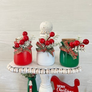 May include: Three small glass jars decorated for Christmas. The jars are painted red, white, and green. Each jar has a plaid ribbon, a sprig of greenery, and red berries. The jars are sitting on a white tiered tray with a white wooden base. The tray is decorated with white beads. The words "Merry Christmas" are written on a red wooden sled.