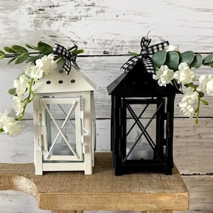 May include: Two small metal lanterns, one white and one black, with a decorative bow and greenery on top. The lanterns are sitting on a wooden stand.